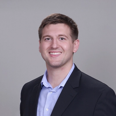 Chiropractor Andrew Plank Smiling In A Suit In Grey Room